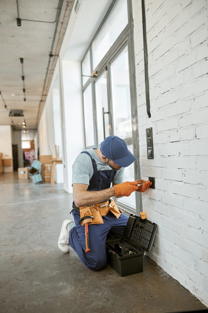 Young man wearing work overalls with tool belt while fixing electric wall socket
