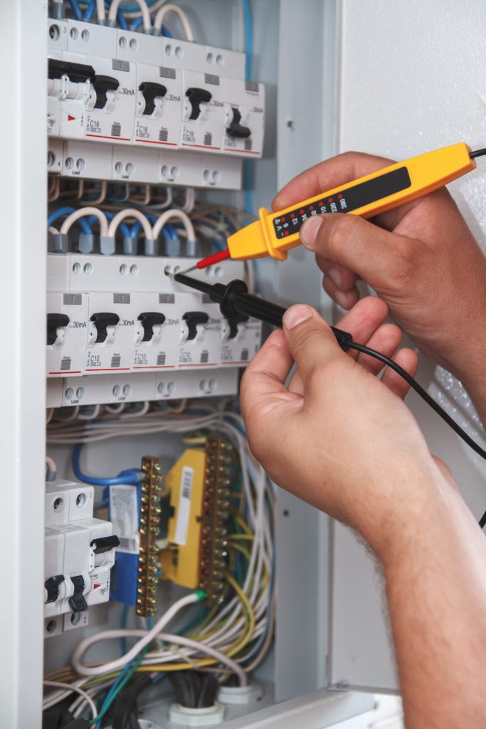 Hands of electrician with multimeter probe at an electrical switchgear cabinet examining fuse box